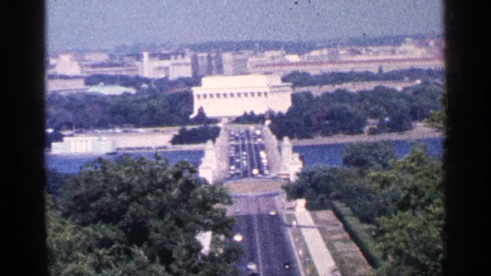 1964 Washington DC 8mm frame showing the Lincoln Memorial from above with water, tree cover, roadway traffic, and city buildings beyond.
