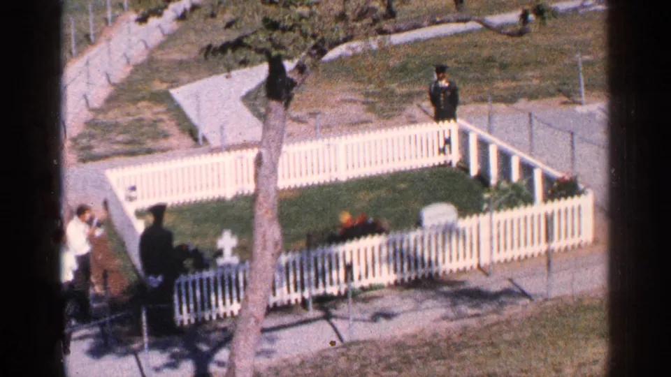 Washington DC 1964 memorial enclosure with a white fence, lawn, markers, visitors, flowers, and a uniformed figure at right.