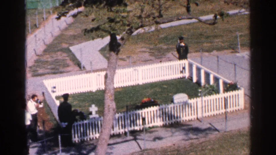 1964 Washington DC home-movie frame showing visitors outside a white fenced memorial enclosure with a tree, lawn, and grave markers.