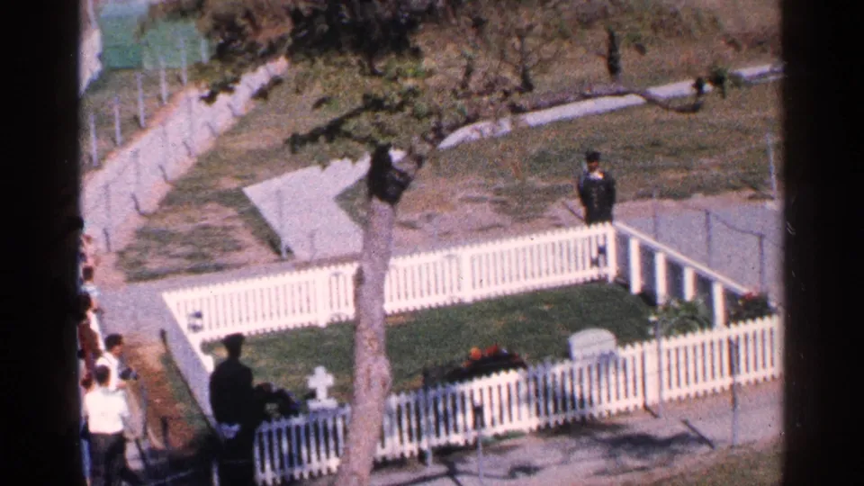 Washington DC 1964 memorial scene with a white fence, small grave markers, flower beds, visitors, and a uniformed figure near the walkway.