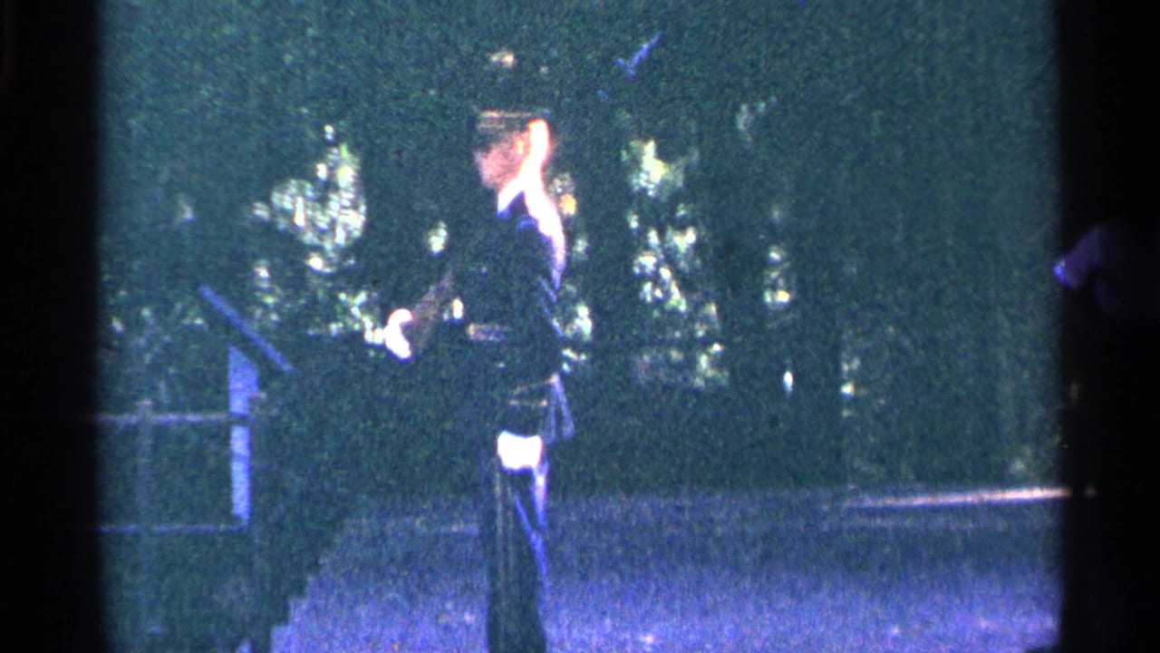 1964 Washington DC home-movie frame showing a uniformed officer saluting near trees and a paved public walkway.