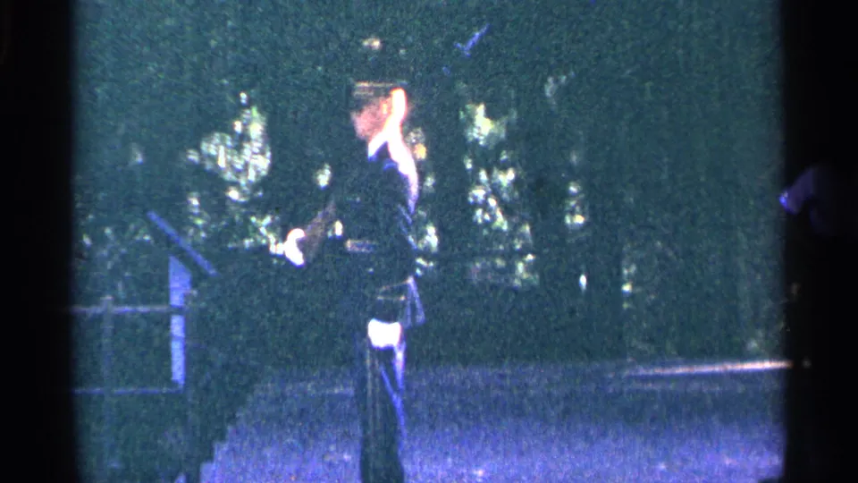 1964 Washington DC home-movie frame showing a uniformed officer saluting near trees and a paved public walkway.