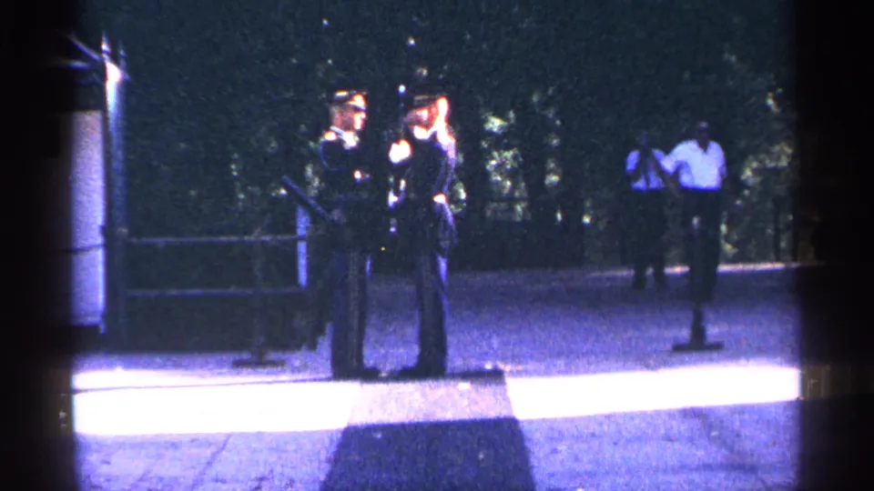 1964 Washington DC frame with a uniformed officer and a civilian adult standing on a paved walkway, with another person in the distance.
