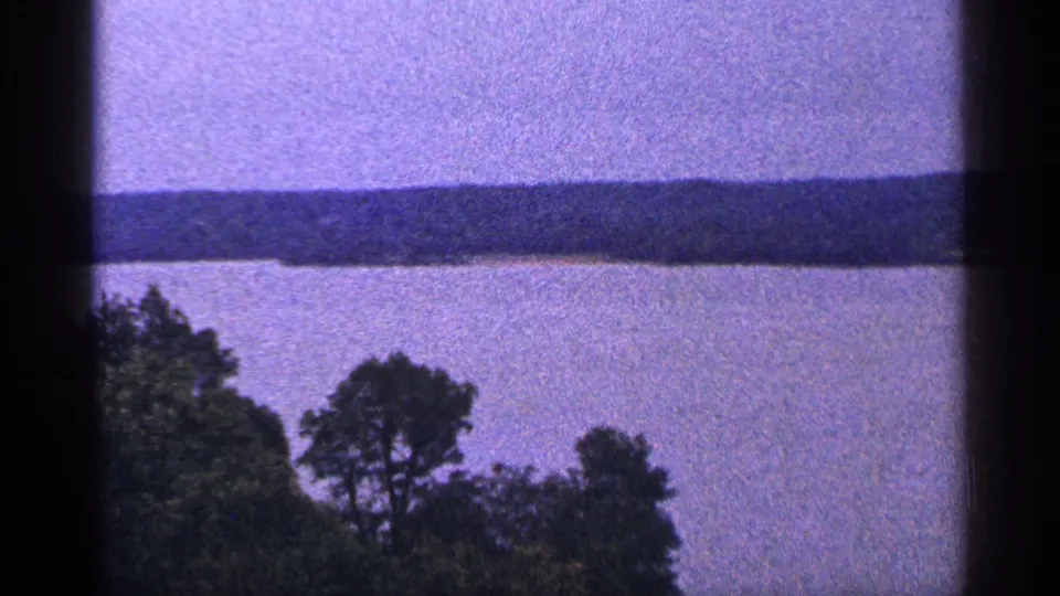 1964 Washington DC 8mm frame showing a calm river, distant wooded shoreline, and dark foreground trees under a color-shifted sky.