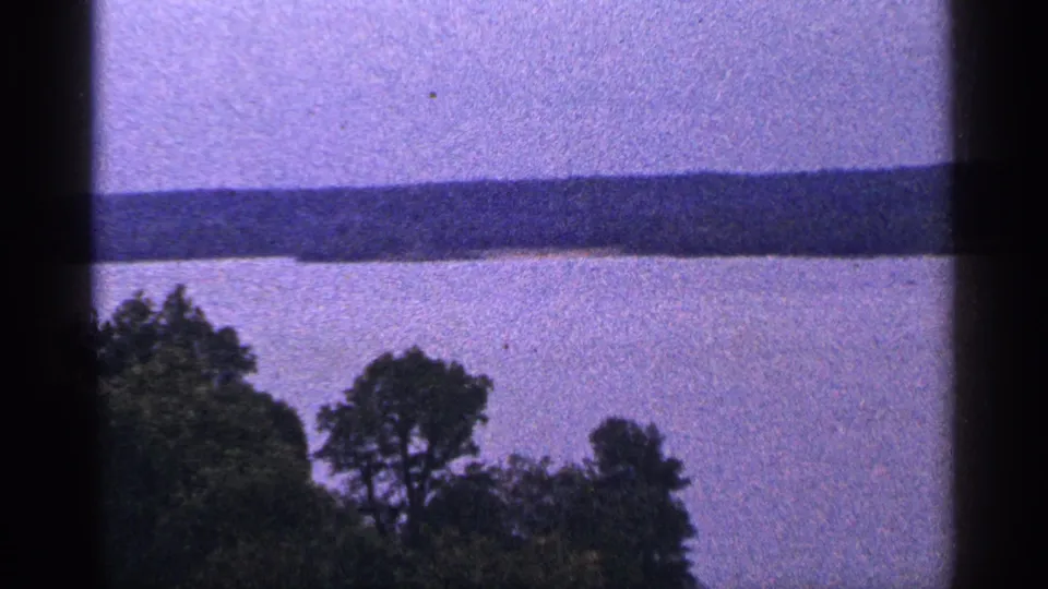 1964 Washington DC 8mm frame continuing the river and wooded shoreline view, with strong grain and dark frame borders visible.