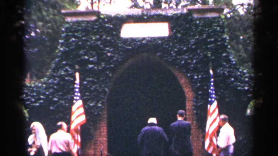 1964 Washington DC home-movie frame showing adults near an ivy-covered masonry arch flanked by two American flags.