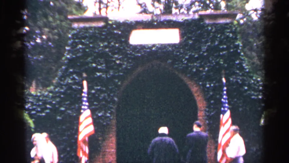 1964 Washington DC 8mm frame of adults moving through a flag-flanked ivy-covered archway at a civic landmark entrance.