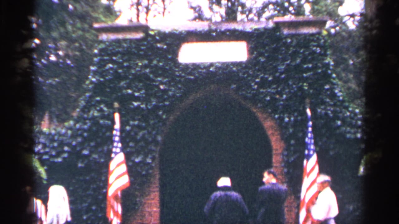 Suited adults near an ivy-covered archway flanked by American flags.