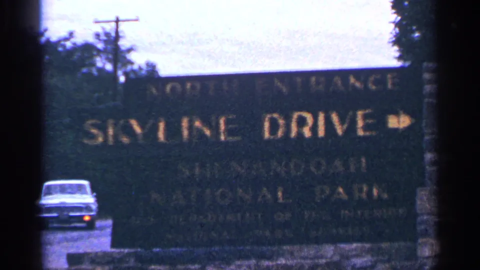 The Shenandoah Skyline Drive north entrance sign fills the frame as a 1960s sedan passes behind it on the road.
