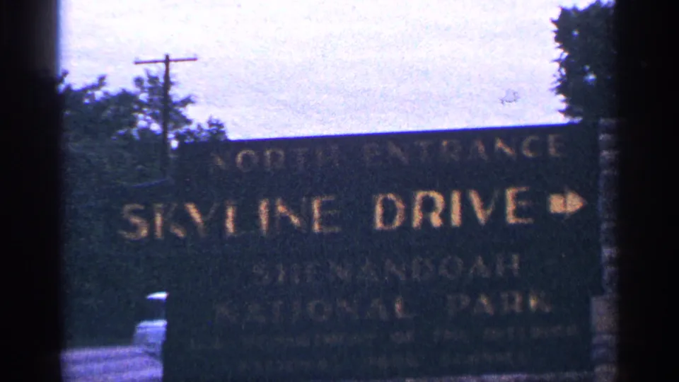 Soft 8mm close view of the Skyline Drive north entrance sign, with gold lettering and roadside trees visible around the sign.