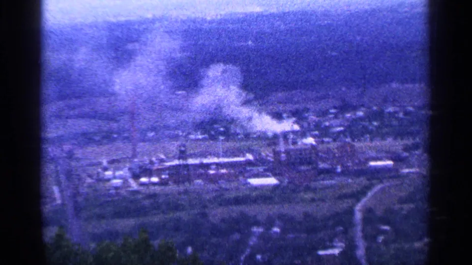 A distant, grainy 8mm view shows buildings and roads below, with smoke or steam rising from an industrial-looking area.