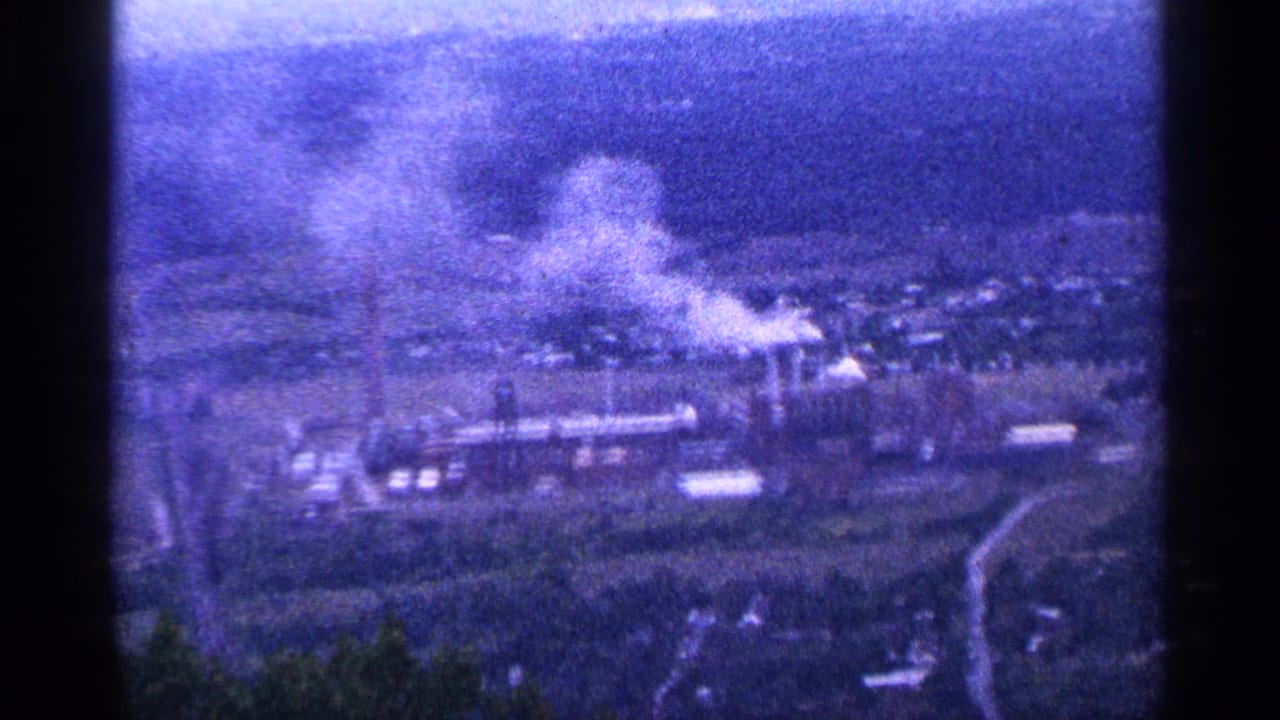 Distant industrial complex in a valley with a white smoke plume and dark mountain backdrop.