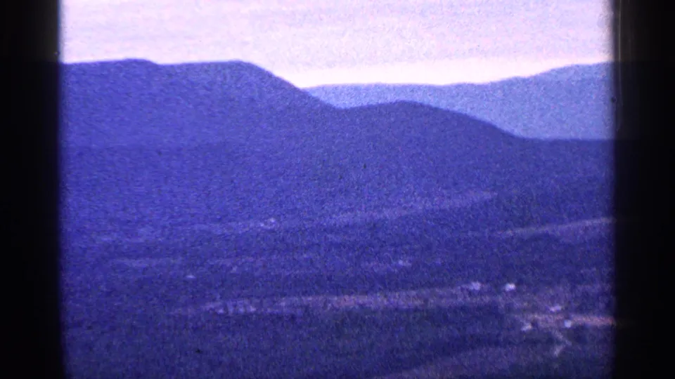 1960s home-movie frame showing blue-toned mountain ridges above a valley with faint roads and small structures.
