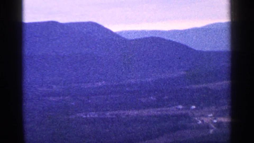 Soft 8mm frame of layered mountains and valley terrain, with small structures and roads visible near the bottom of the view.
