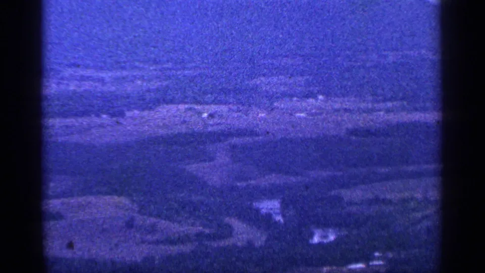 1964 8mm frame showing a blue-purple hazy mountain landscape with ridges, valley terrain, and rocky foreground from an overlook.