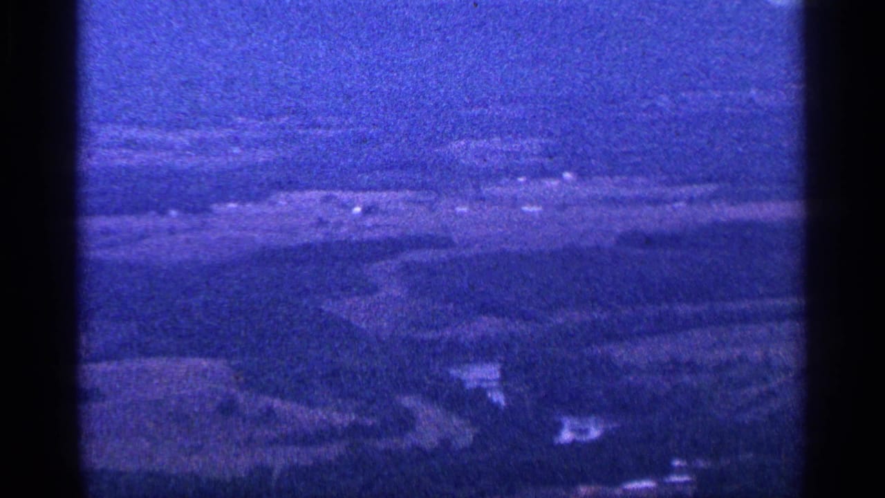 1964 8mm home-movie frame showing hazy farmland and distant hills under a cloudy sky, with a strong blue cast and visible gate edges.