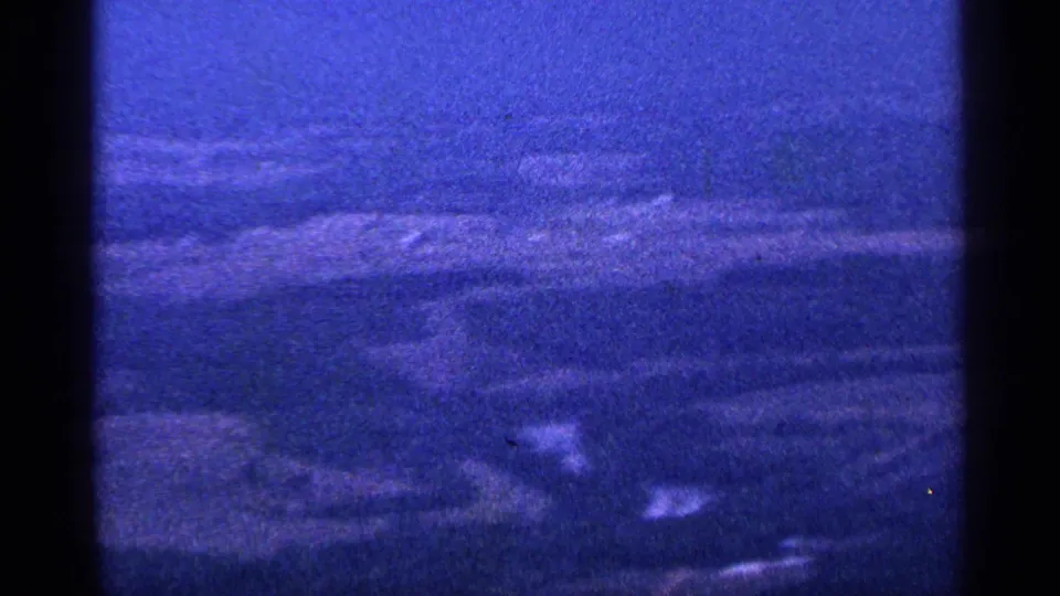 Blue-tinted 1964 8mm frame of rural fields and a hazy hill line, likely filmed from a moving aircraft or high vantage point.