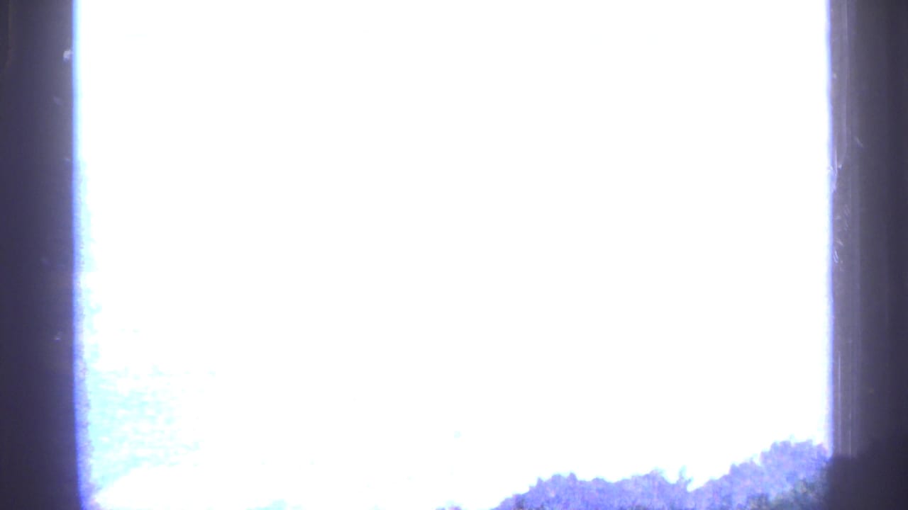 1964 8mm home-movie frame with heavy overexposure, dark gate edge, and a faint tree-covered hillside along the lower edge.