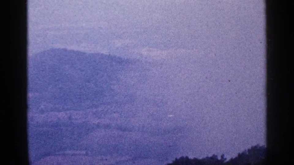 1964 8mm mountain vista with blue-purple haze, distant ridges, lower tree line, and a dark frame border at left.