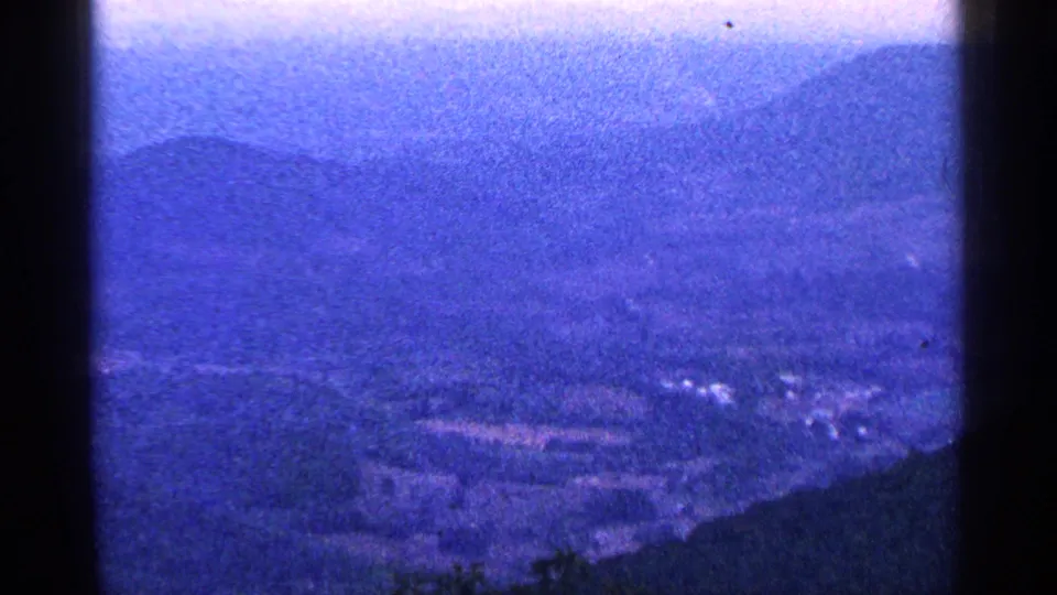 Blue-tinted 8mm view from the overlook toward hazy ridges, valley land, and small pale structures.