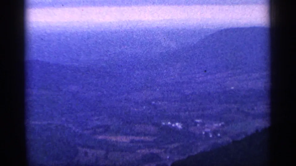 Grainy 8mm frame showing a hazy valley and layered hills from a high overlook perspective.