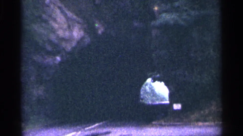 A paved road leads toward a tunnel opening below a rocky wooded embankment in 1964 West Virginia travel footage.