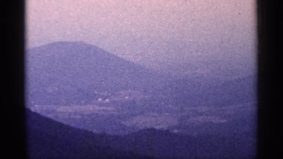 1964 West Virginia 8mm frame showing a broad hazy valley and layered purple-blue mountain slopes beneath a washed-out sky.