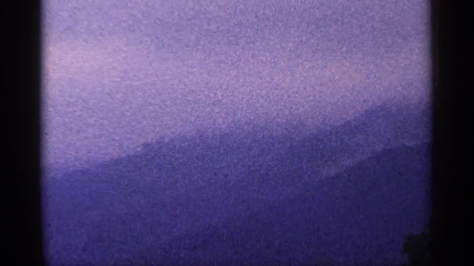 1964 West Virginia 8mm frame showing a soft purple mountain ridge beneath a pale hazy sky, with heavy grain and dark frame borders.