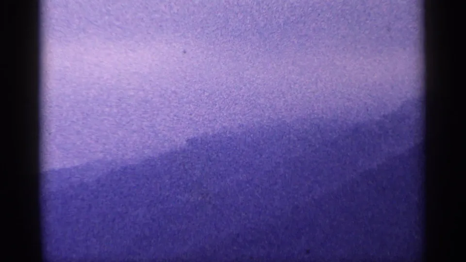 1964 West Virginia 8mm frame with soft purple hills, pale sky, heavy grain, and subdued contrast across the landscape.