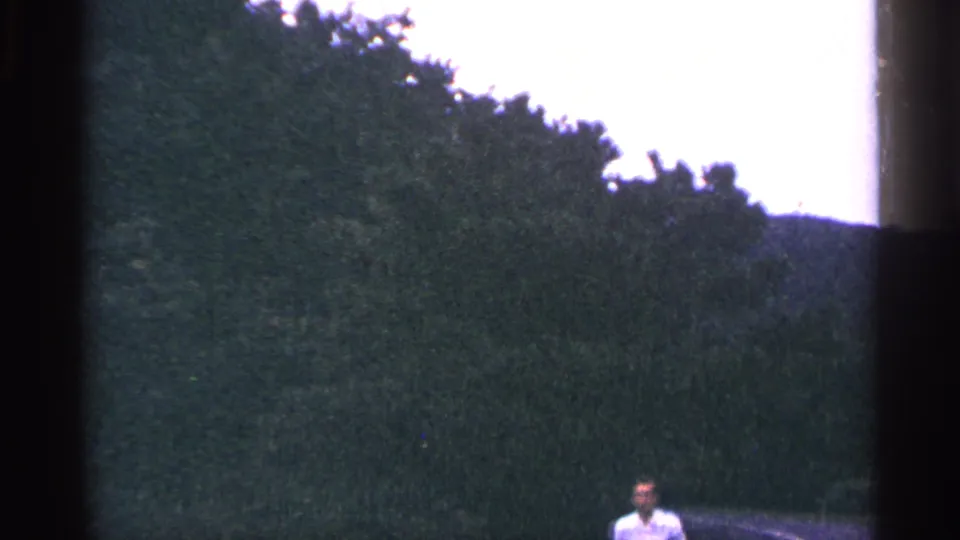 West Virginia 8mm frame showing an adult man in a light shirt near a curved road, with dense trees and hills behind him.