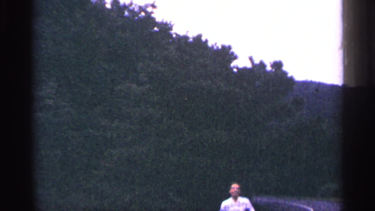 In 1964 West Virginia 8mm footage, a man in a light shirt appears near a roadside with dense trees and part of a parked car.