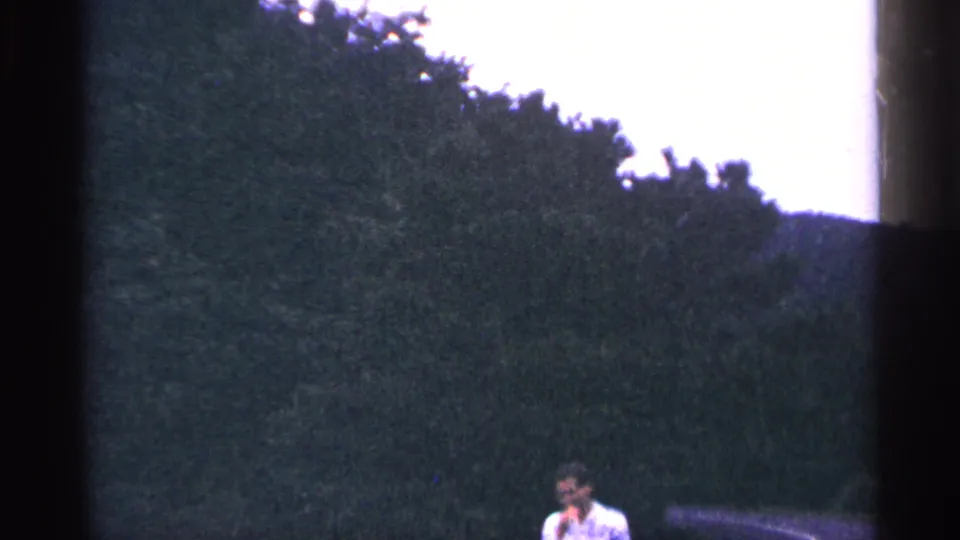 The man remains partly visible near a car against dark green trees in blue-tinted 1964 West Virginia home-movie footage.