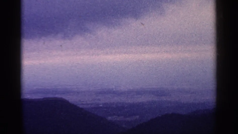 1964 8mm frame of West Virginia mountain silhouettes beneath a softly clouded purple-blue sky.