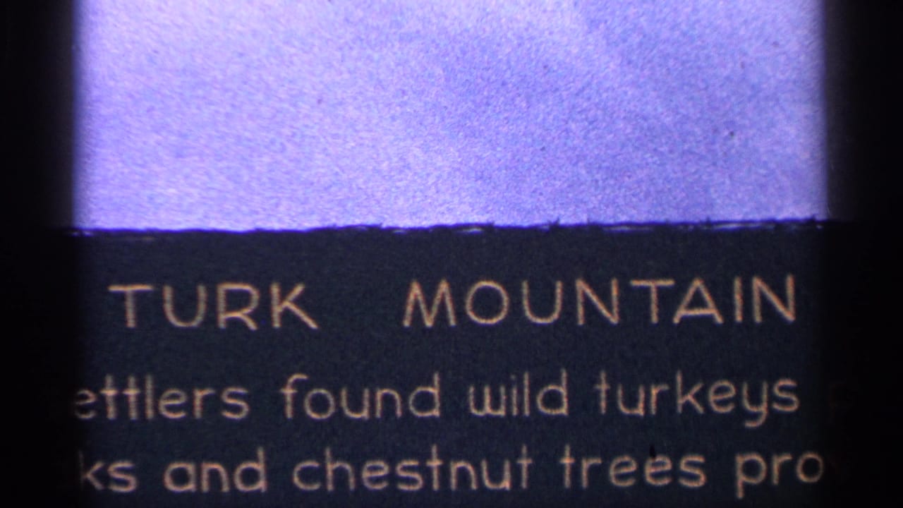 1964 West Virginia 8mm frame showing a partial TURK MOUNTAIN interpretive text panel next to a purple-toned mountain view.