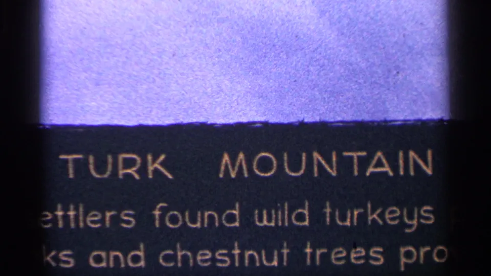 1964 West Virginia 8mm frame showing a partial TURK MOUNTAIN interpretive text panel next to a purple-toned mountain view.