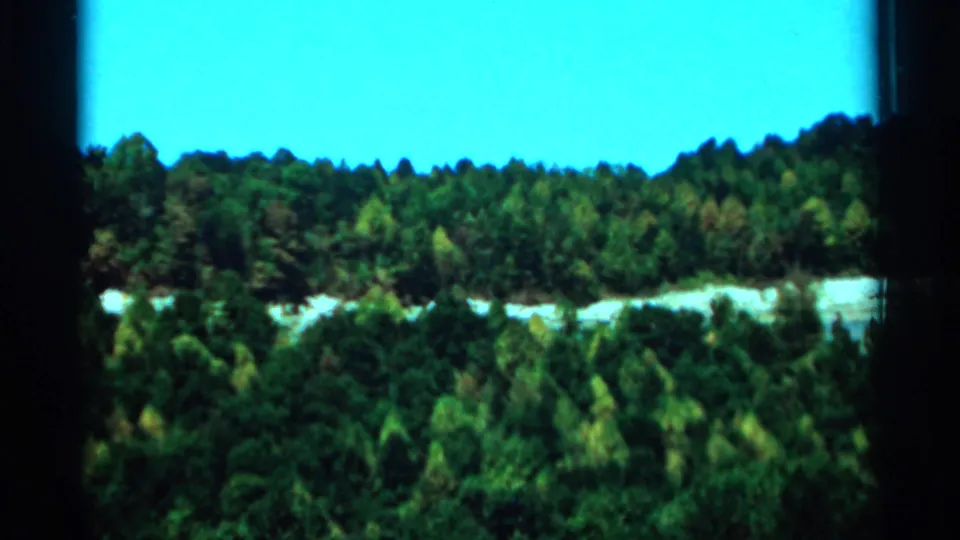 1964 West Virginia home-movie frame showing a wide elevated forest view under bright blue sky with a pale river or path.