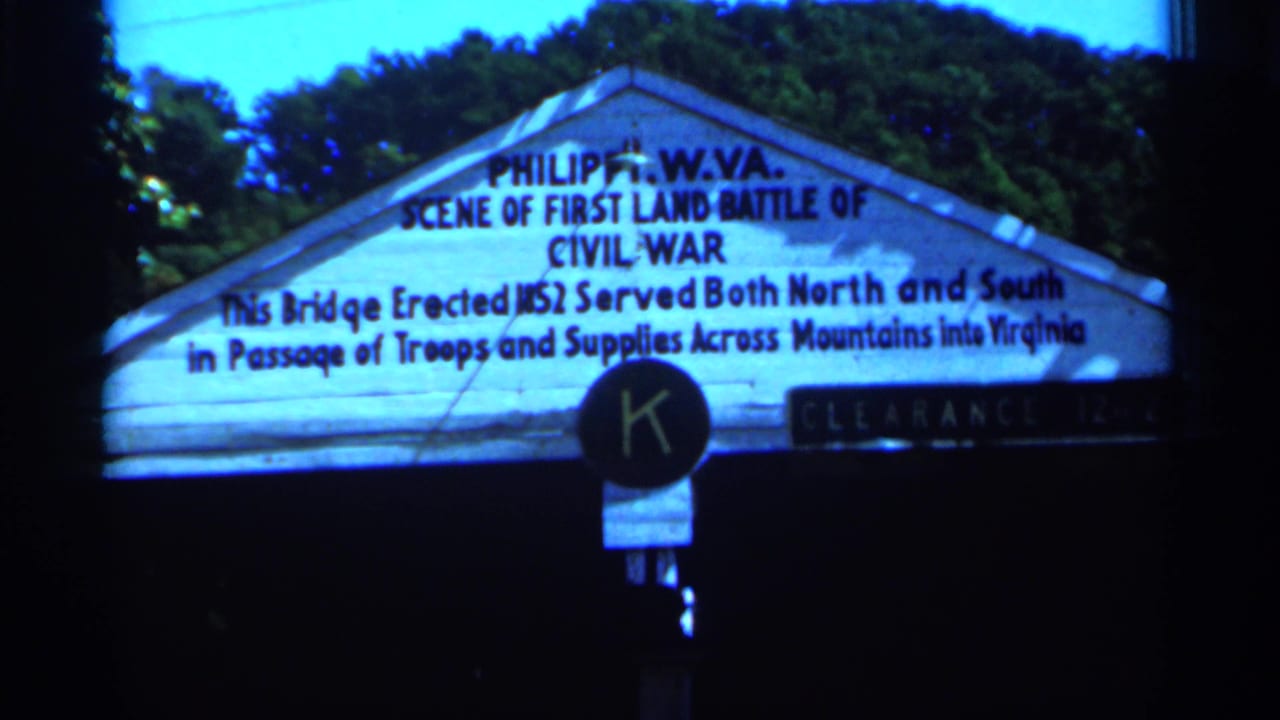 1964 West Virginia 8mm frame showing a white Philippi Civil War marker sign with surrounding trees and dark frame edges.