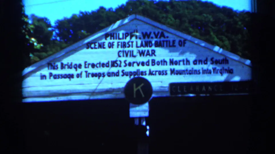 1964 West Virginia 8mm frame showing a white Philippi Civil War marker sign with surrounding trees and dark frame edges.