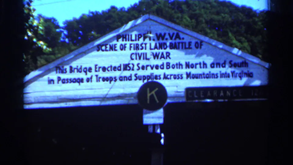 1964 West Virginia 8mm frame showing the Philippi Civil War roadside marker, with dense trees and dark film edges.