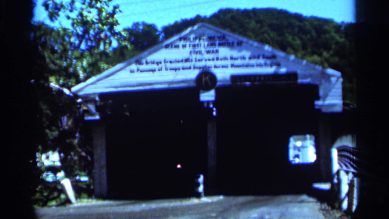 1964 8mm frame of a white covered bridge in West Virginia, with Civil War text across the gable and trees behind it.