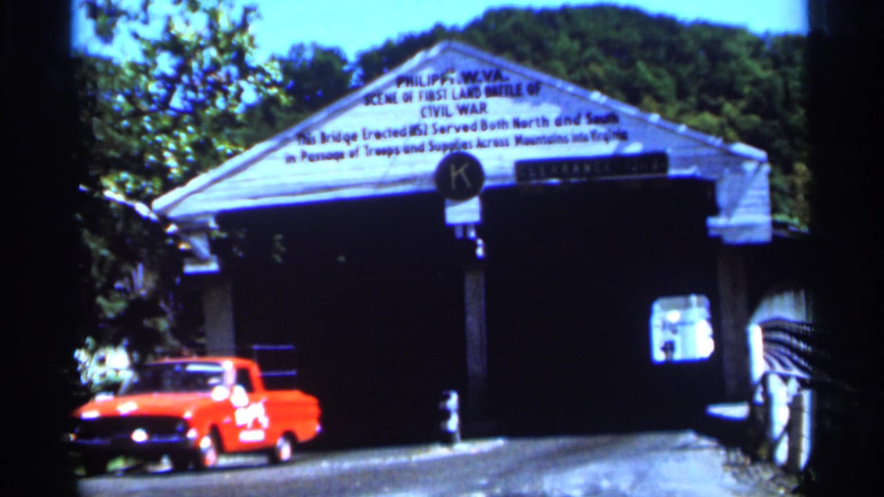 Red vintage pickup drives toward a white West Virginia covered bridge with Civil War text and a clearance sign visible above.