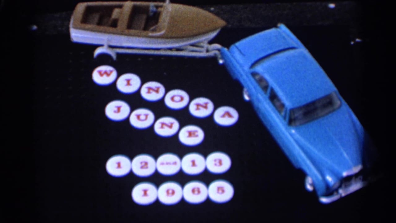 Toy blue car towing a small boat beside round tiles spelling Winona June 12-13 1965.
