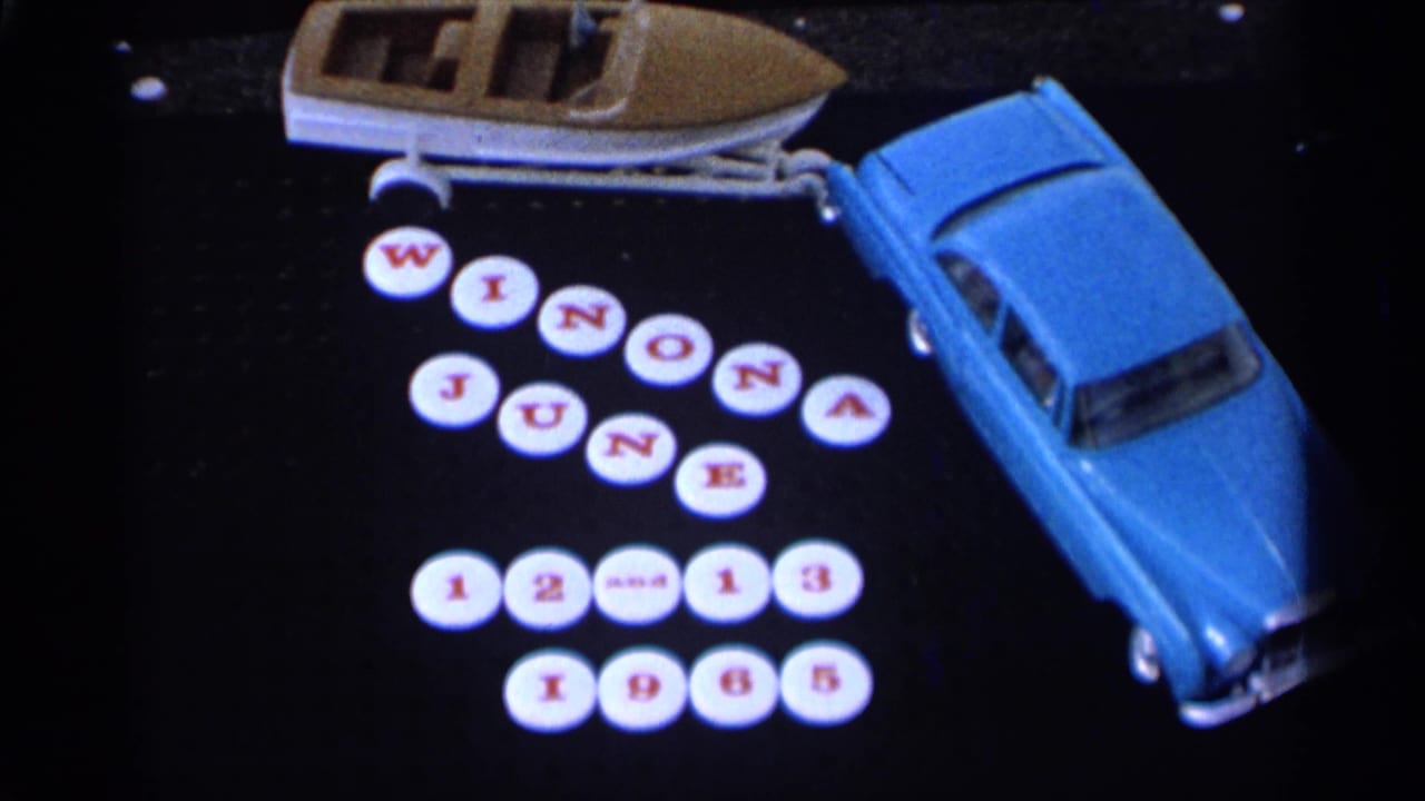 Blue toy car towing a small boat on a dark surface, with white round tiles forming Winona June 12-13 1965.