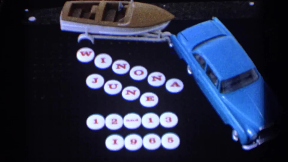 Close 8mm view of a blue toy car and boat beside round tile letters marking Winona, June 12-13, 1965.