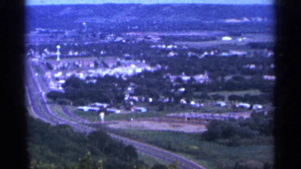 Soft 1960s 8mm aerial or elevated view of a town area with roads, scattered buildings, open ground, and distant horizon.