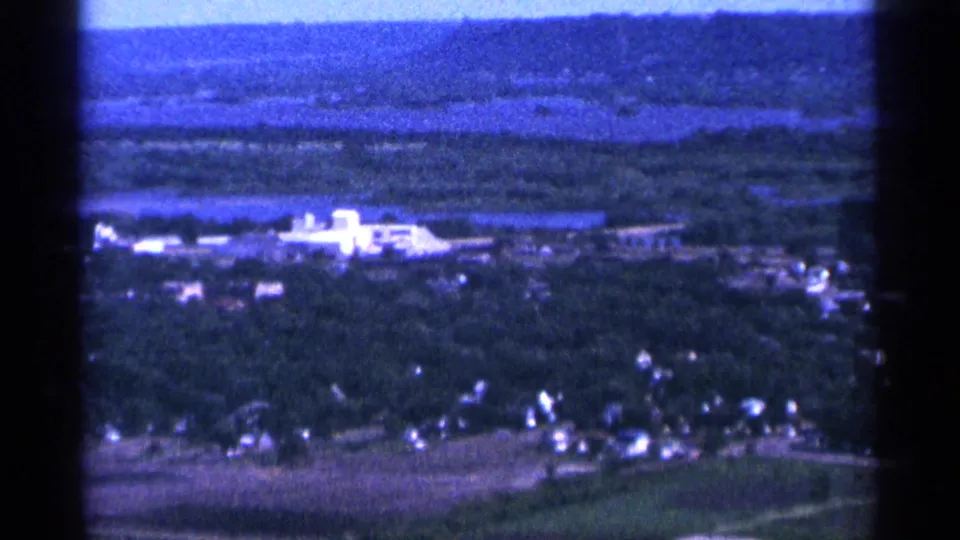 1965 Duluth-area frame showing pale low buildings, wooded foreground, fields, and distant water or lowland.