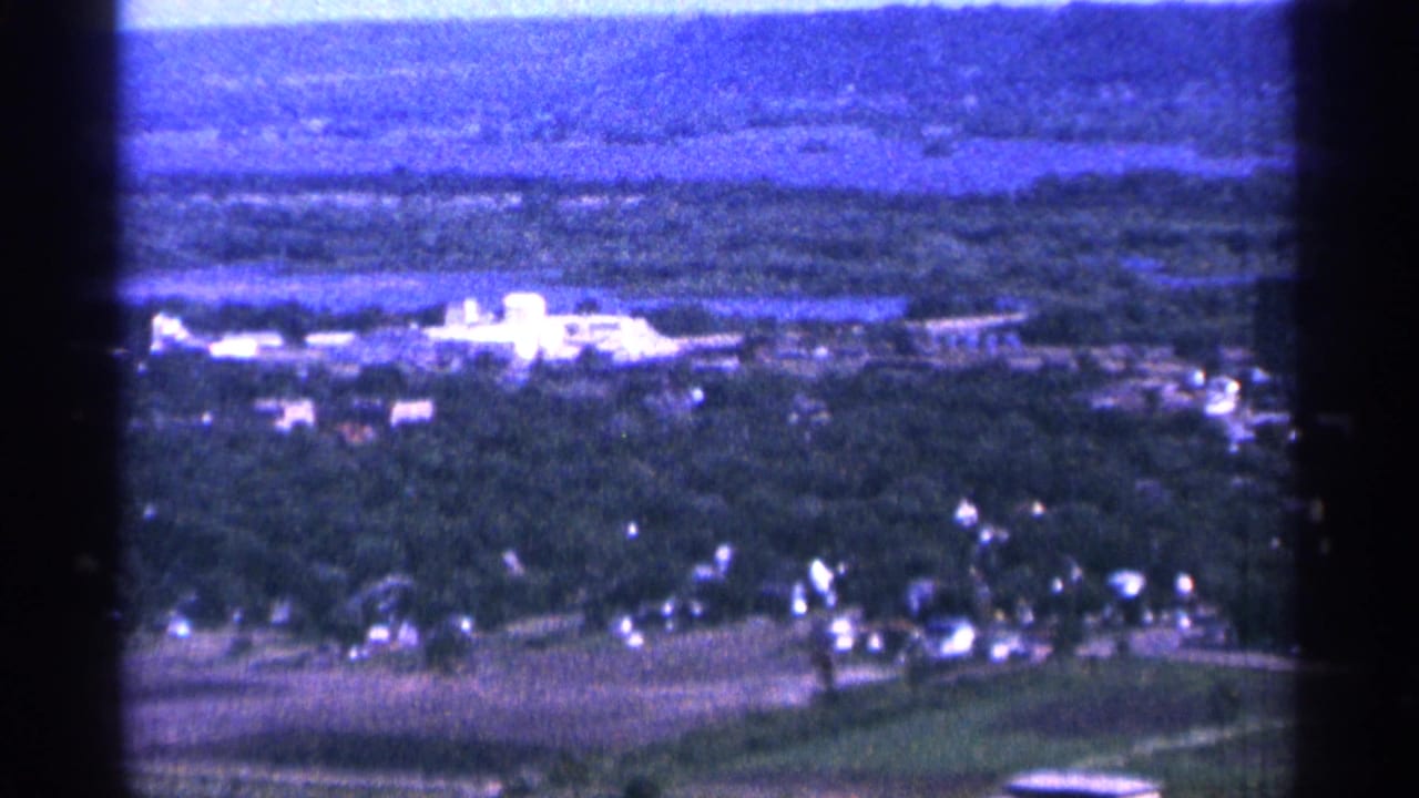 1965 Duluth, Minnesota aerial 8mm frame showing a wooded community, open fields, scattered houses, and a bright white building near a body of water.