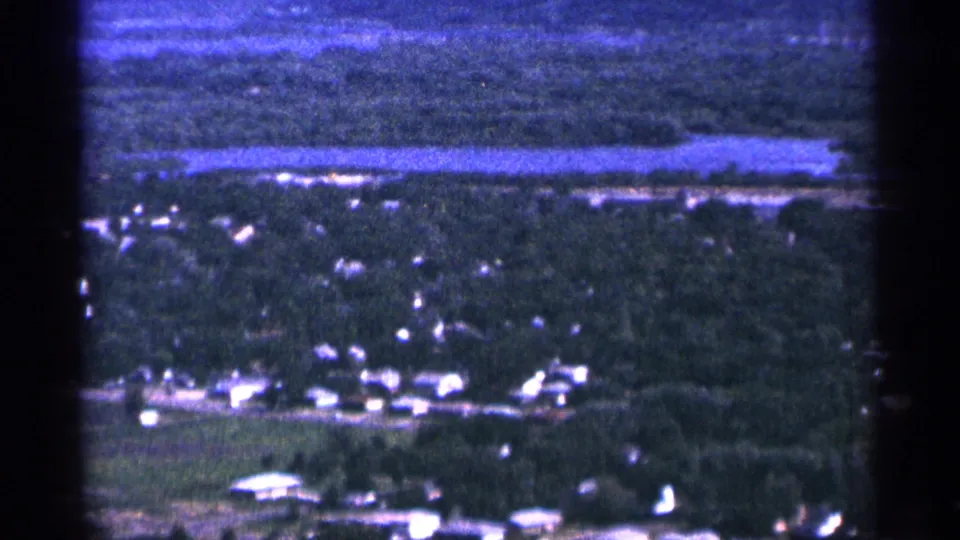1965 Duluth, Minnesota aerial frame showing tree cover, scattered buildings, and a long horizontal body of water beyond the town.