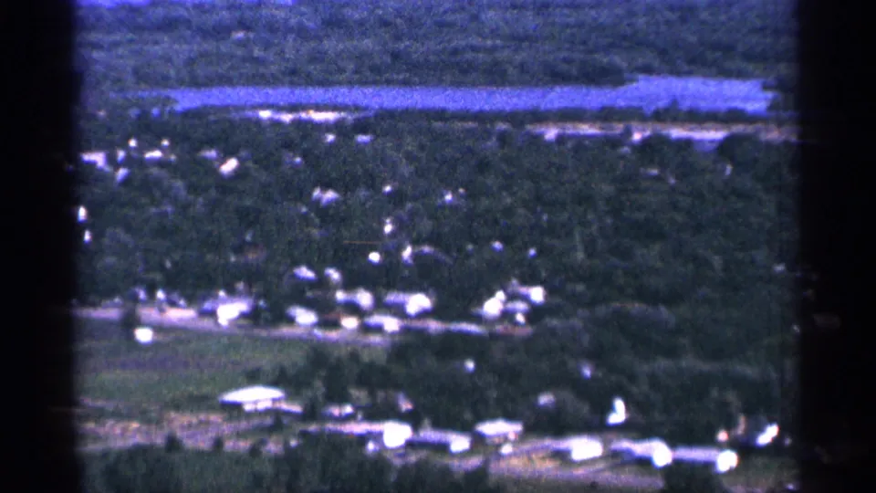 1965 Duluth, Minnesota aerial 8mm view of houses and farm-like clearings among trees, with water stretching across the upper frame.