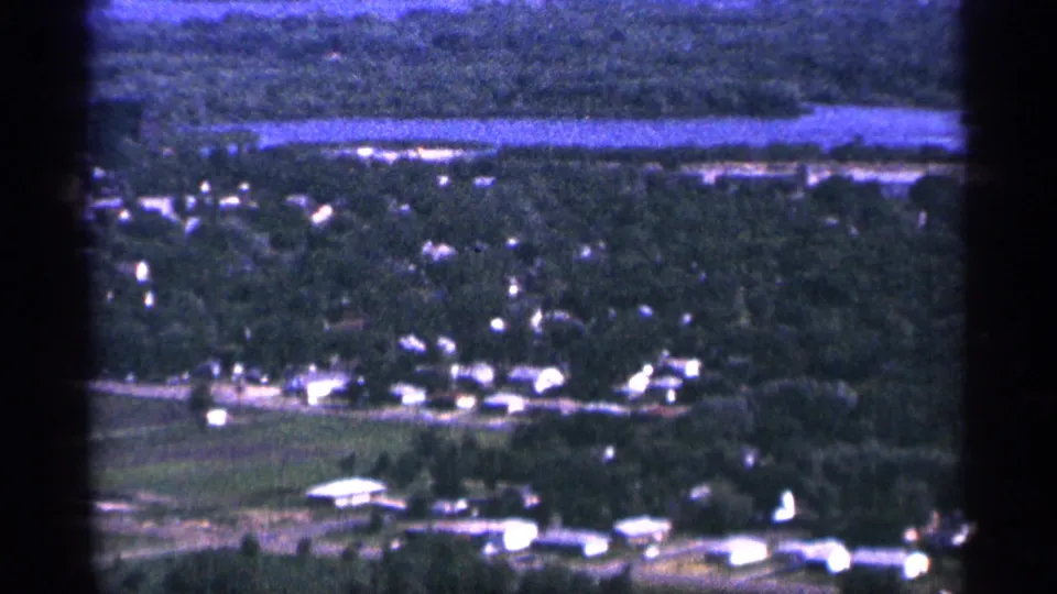 1965 Duluth, Minnesota aerial frame showing a small wooded settlement, bright rooftops, open ground, and a distant strip of water.